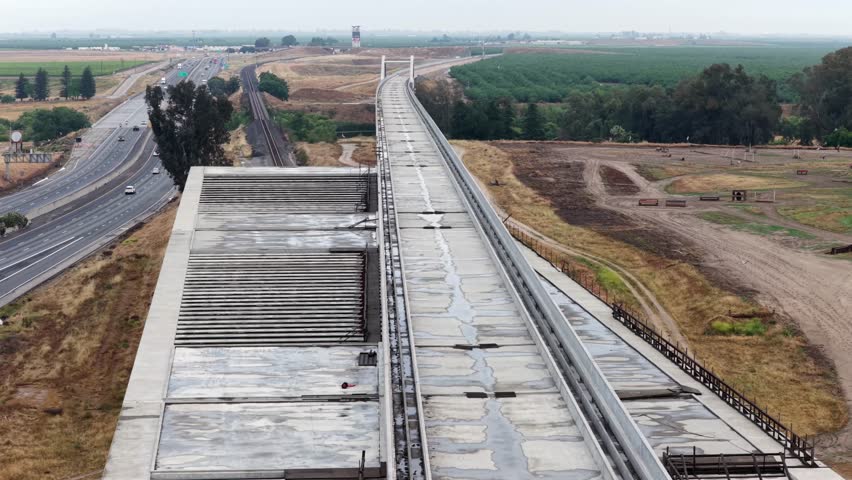 California’s concrete high-speed rail viaduct curves through Fresno’s flat landscape, cars travel adjacent highway past green orchards and dirt construction site below. Aerial drone wide shot