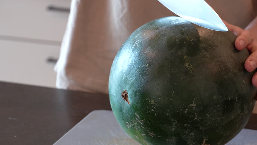 Man cuts a ripe seedless watermelon in half with a kitchen knife and breaks it open to reveal juicy red flesh. Fresh summer fruit, close-up food preparation