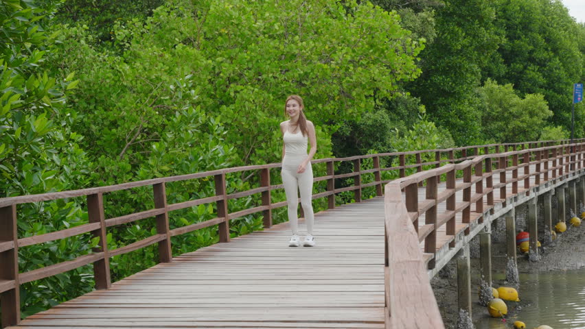 Active female athlete running outdoors on wooden boardwalk trail, smiling Asian woman in white sportswear doing cardio workout for healthy lifestyle in beautiful green nature park.