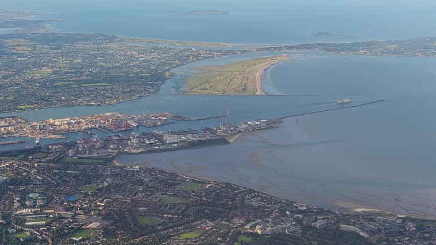 Aerial view overlooking the River Liffey estuary and Dublin Port industrial zone in Ireland. Features clear visibility of the iconic red-and-white Poolbeg stacks, tidal mudflats, Bull Island beach,