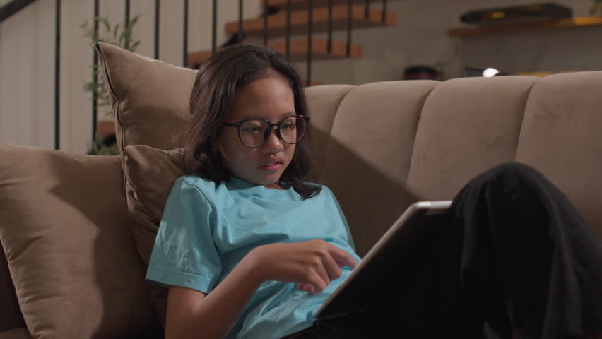 An Asian girl wearing glasses reads from a tablet while leaning comfortably on a living room sofa, capturing home learning, digital education, childhood literacy, and modern family life.