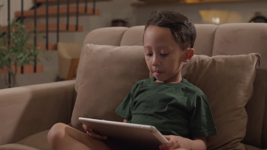 Little boy using a tablet while sitting on a living room sofa, capturing children
