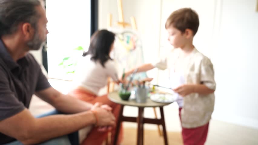 The family—father, mother, and son—are doing a family activity together. The father is teaching his son to draw and paint on a canvas at home.