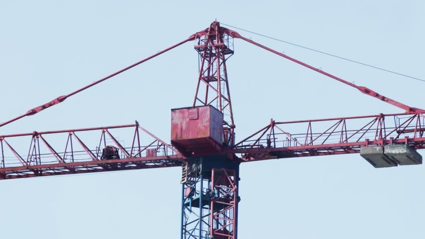 View of upper section of powerful tower crane, red jib and counter-jib extend from central mast, blue base structure visible, industrial equipment for lifting heavy loads, construction site machinery