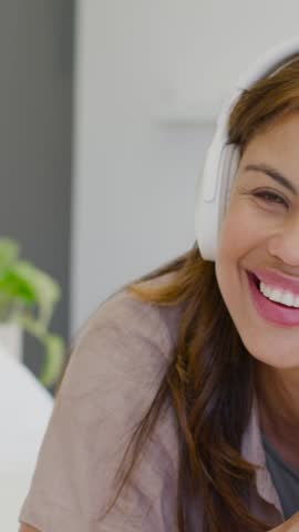 Vertical video: Listening to music, woman pressing white headphones on bed, wearing pink smartwatch. Bedroom, relaxed, cozy, sunlit, casual, plant, vertical video