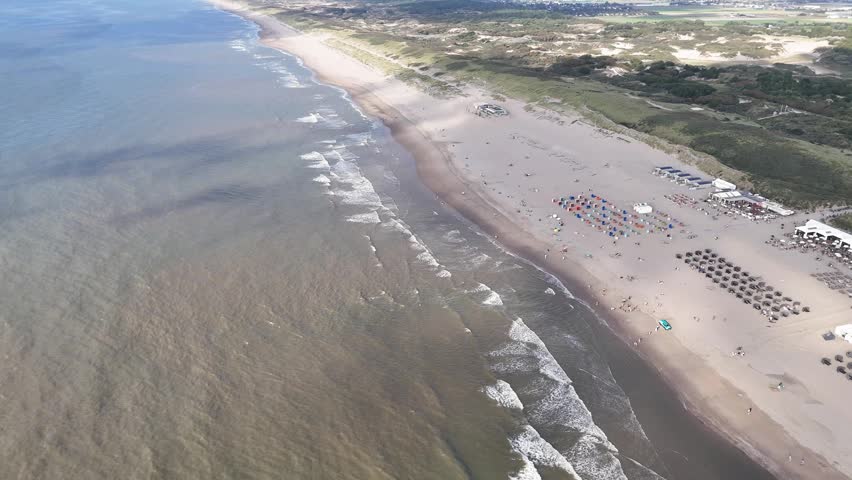 An aerial footage of the North Sea waves rolling along the sandy coast of Noordwijk city in the Netherlands on a sunny day