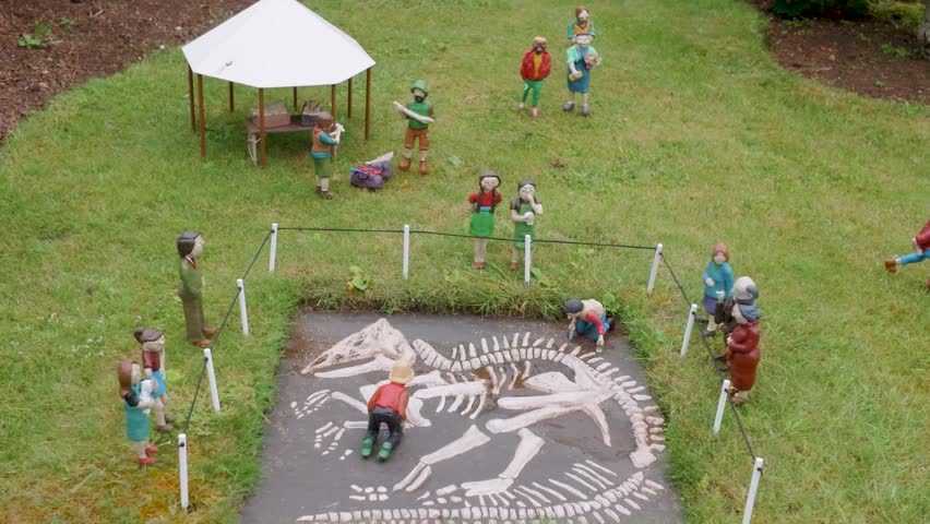 Model village dinosaur dig scene with miniature figures around exposed fossil skeleton in fenced pit; camera orbits and reverses, ideal for museum, education, and prehistory themes.