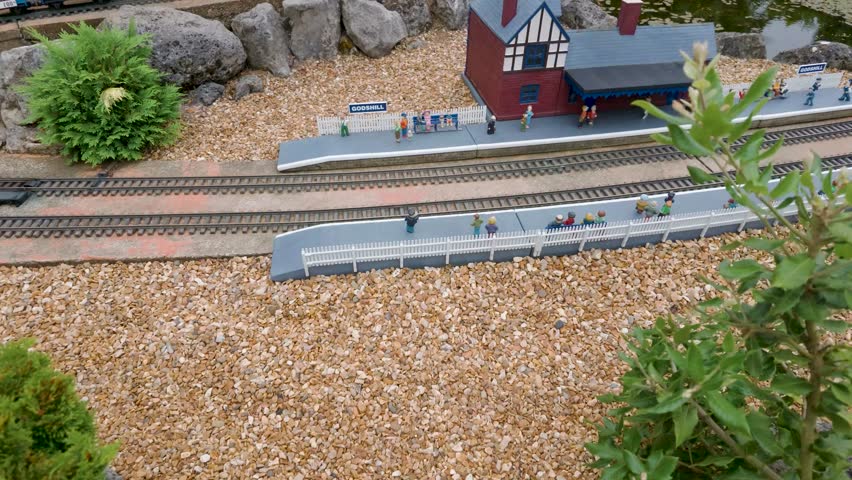 Model train arrives at Godshill Model Village station, pulling in and stopping as dinosaur-themed carriages pass miniature platforms, tiny figures and buildings in UK attraction.