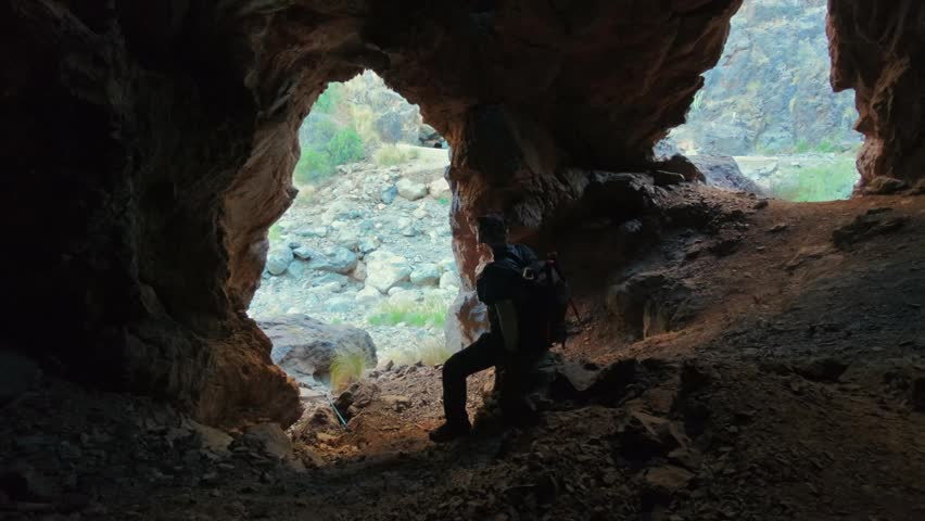 Adventurous man with a backpack resting at the mouth of a cave. Explorer contemplating the landscape from inside the grotto