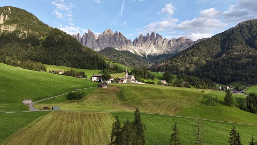 Wide drone panorama of Santa Maddalena Church, rural fields and forested valley beneath the Odle Mountains in Val di Funes, Italy.