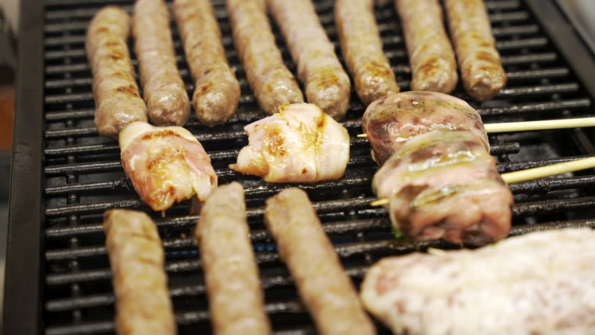 Rows of sausages and skewered meat cook on a hot barbecue grill inside a food store.
