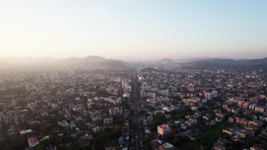 An aerial view of a dense city with a long road stretching into the distance, surrounded by hazy hills at sunset