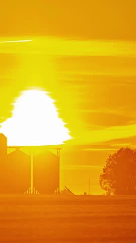 View of grain silos silhouetted against a vibrant golden sunset sky in the rural landscape of Latvia.