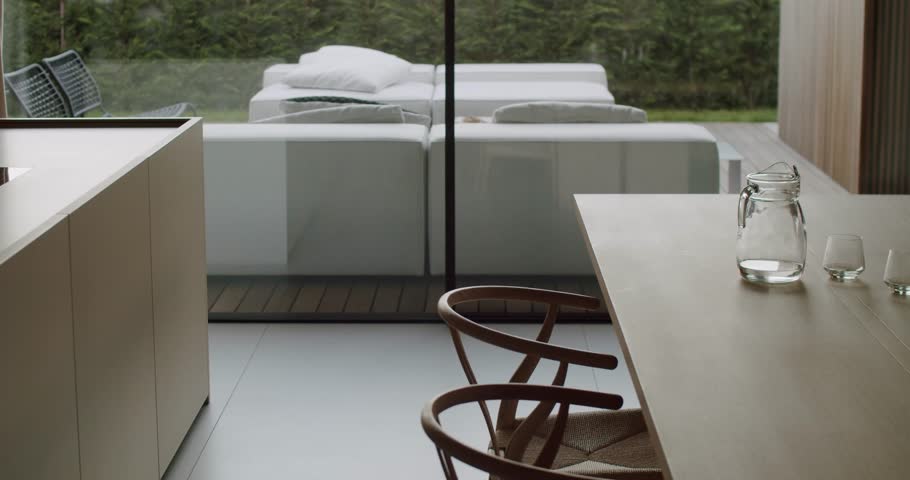 Modern dining room with wooden table, chairs, and water jug. Minimalist interior design with a view of the patio and garden through large windows. Natural light illuminates the scene, creating a calm