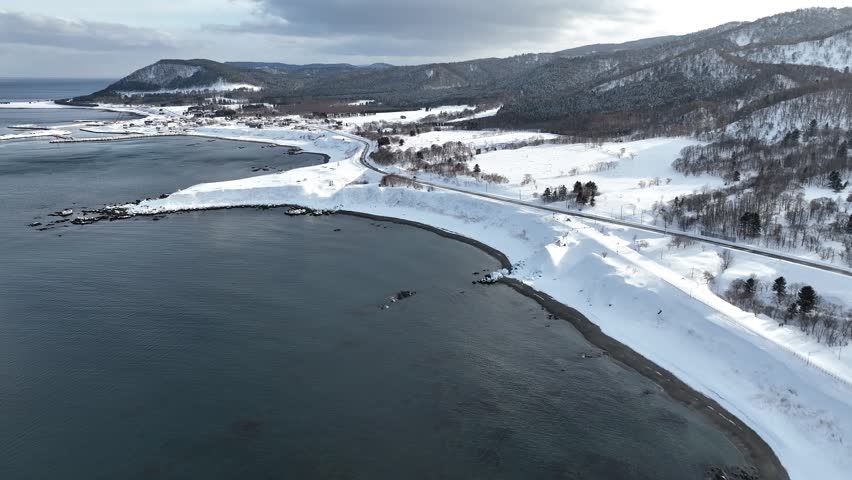 Aerial footage of Cape Kitami Kamui on the Sea of Okhotsk coast in Hokkaido, Japan. Filmed from the Esashi side on a calm winter afternoon, the landscape is almost entirely covered in snow. The black-and-white Cape Kitami Kamui Lighthouse stands above dramatic coastal cliffs, while traces of the former Okhotsk Main Line railway can still be seen beneath the snow. The nearby former coastal road remains closed during winter, making aerial imagery one of the few ways to appreciate this remote landscape.