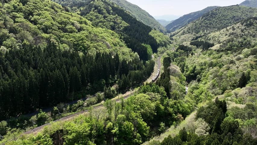 Aerial footage of an E721 Series local train arriving and departing Omoshiroyama-Kōgen Station on the JR East Senzan Line. Captured on a clear early summer day, the video follows the four-car commuter train through a beautiful mountain gorge surrounded by forests, waterfalls, and the historic Senzan Tunnel portal. One of the most scenic railway sections connecting Yamagata and Sendai.