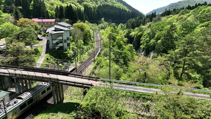 Aerial footage of an E721 Series local train arriving and departing Omoshiroyama-Kōgen Station on the JR East Senzan Line. Captured on a clear early summer day, the video follows the four-car commuter train through a beautiful mountain gorge surrounded by forests, waterfalls, and the historic Senzan Tunnel portal. One of the most scenic railway sections connecting Yamagata and Sendai.