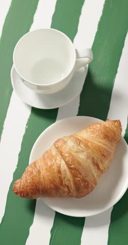 freshly brewed black coffee being poured from a coffee pot into white cup, served with buttery croissant on plate on rustic white and green striped wooden cafe table. Cozy breakfast scene with fresh pastry and hot coffee, perfect for cafe, bakery, morning routine, brunch, hospitality, and food advertising concepts. Premium 4K food video
