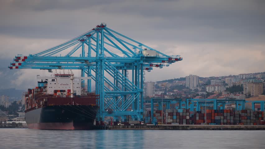 View of a container ship docked at the Adriatic Gate Container Terminal with blue gantry cranes, stacked shipping containers, and city buildings, Rijeka, Primorsko-goranska zupanija, Croatia.