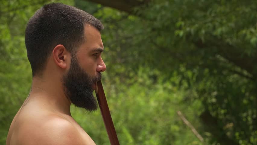 Bearded man playing a traditional kaval flute. Long pipe wooden wind instrument performance in a green forest.