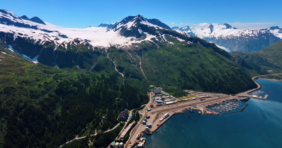 Piece of urban area with buildings, roads and berths with boats surrounded by green snow-capped mountains and blue waterscape. Aerial perspective on the scenery in Alaska.