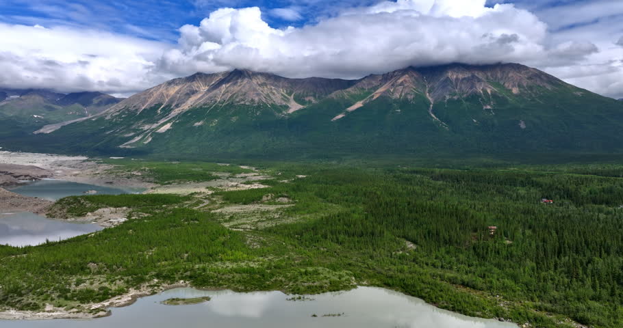Rising over the river in the valley with pine tree woods. White fluffy cloudscape covers the stunning mountains in Alaska.