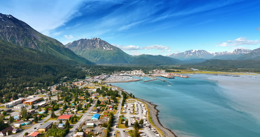 Flight over the town in Alaska scattered on the waterfront of the turquoise river. Pine tree woods and spectacular mountains surround the urban area.