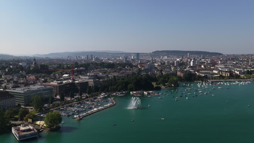A drone view of Zurich city on the edge of Lake Zurich, with a fountain and moored boats under a sunny sky in Switzerland