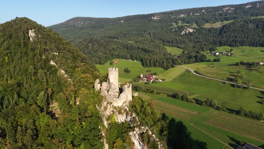 A drone view of Neu-Falkenstein Castle with a tall tower with flag sits atop a rocky hill, surrounded by a green, mountainous landscape in Switzerland