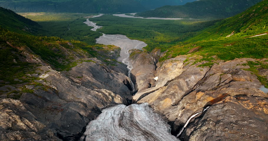 Flight over the glacier in the high mountains. Approaching the river in the beautiful valley at the mountain foot. Alaska scenery.