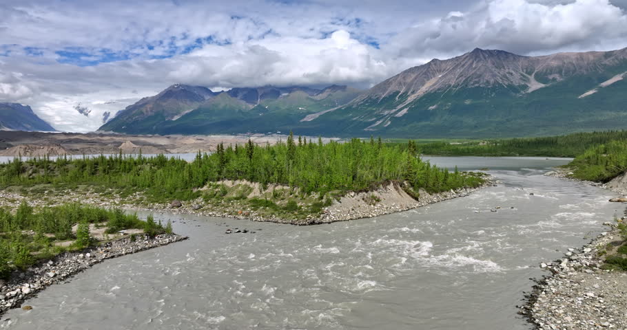 Flight over the fast river approaching the pine tree wood. Beautiful mountains covered with white cloudscape at backdrop. Alaska sceneries.