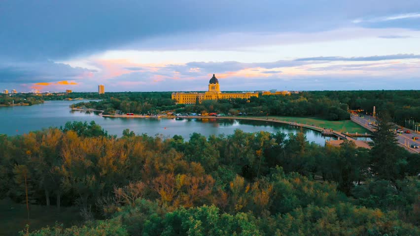 Golden light illuminating the Saskatchewan Legislative Building across Wascana Lake, framed by autumn trees under a dramatic twilight sky in Regina