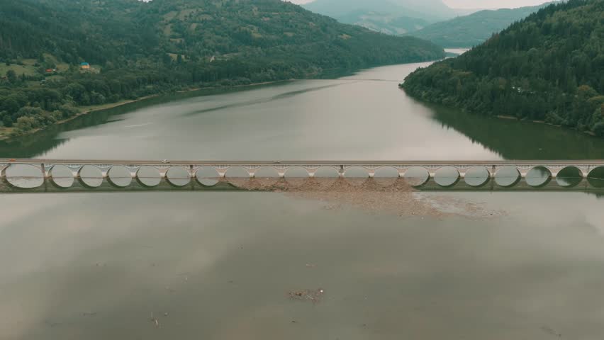 An aerial view of Bicaz Lake Bridge crossing calm waters with forested hills and mountains in the background