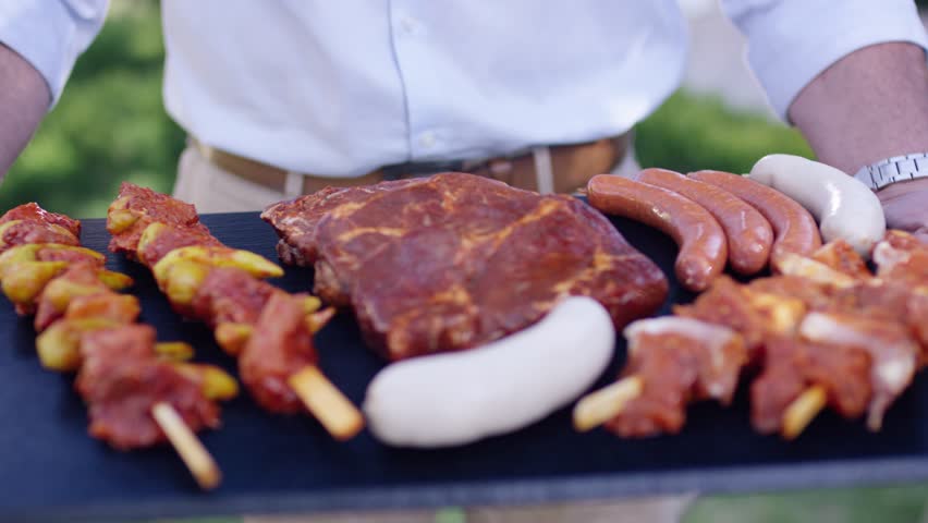 Man in a light blue shirt carrying a slate tray with raw meat skewers steaks and sausages outdoors.