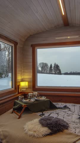 View of cozy cabin interior with wooden walls, soft blankets, and a breakfast tray overlooking a serene snowy landscape in Latvia.