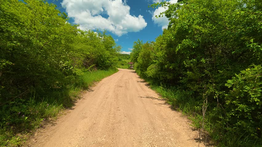 Dirt road winds through lush greenery beneath a bright sky. Silver car rests beside the path, shaded by trees. Location: Vrmdza lake hike (Vrmdzansko jezero), Mount Rtanj area. Balkans, Serbia