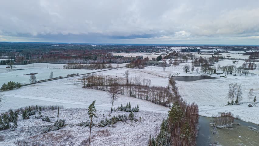 Aerial view of a snowy rural landscape featuring fields, a small pond, and a road under an overcast sky in Latvia.