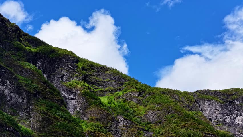 Aerial view of the steep, rocky mountainside in Flam covered in lush green vegetation under a bright blue sky with white clouds in Flam, Vestland, Norway.