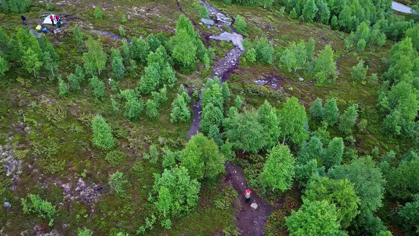 Aerial view of a small stream winding through lush green trees and mossy terrain in the mountains of Flam, with a tent and hikers visible in Flam, Vestland, Norway.