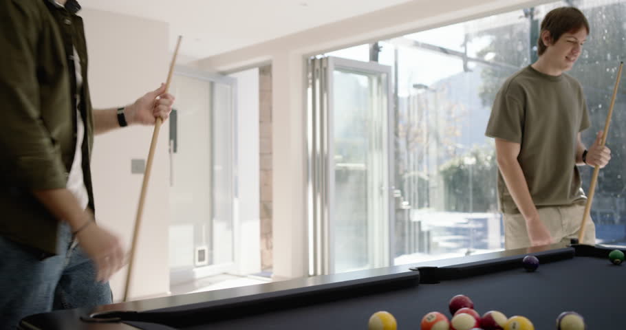 Diverse male friends gathering at pool table, one placing cue ball, one shooting for fun. Billiards, companions, social, recreation, sunlit, patio, modern