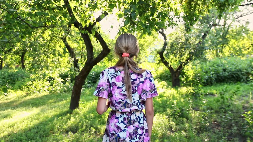 Back view of teenage girl running away into green sunny garden. Young female in colorful dress enjoying summer day in park, out of focus.