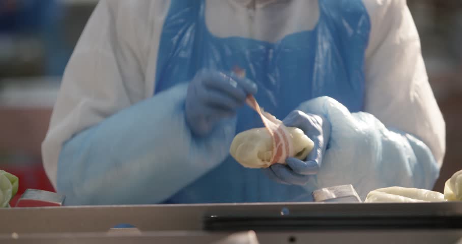 Person in protective clothing wrapping a cabbage roll with bacon in a food production facility.