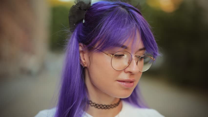Young woman with purple hair and glasses adjusting hair outdoors. Stylish female with colorful dyed hair smiling directly at the camera on a city street in summer