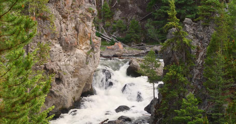 A medium shot focusing on churning white water rapids as they surge past a prominent rocky cliff face and small evergreen trees.