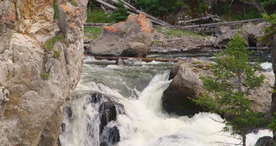 A tight, close-up shot capturing a river dropping sharply over a rocky ledge, with lichen-covered boulders and a small pine tree on the bank.