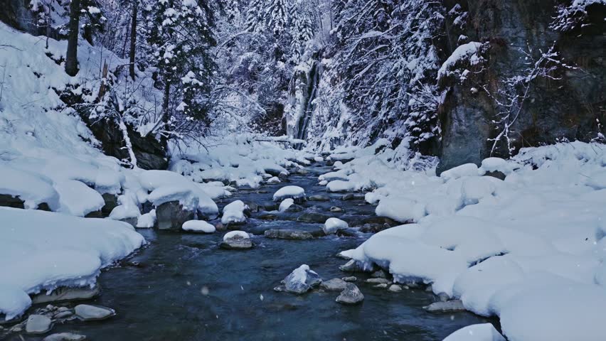 Peaceful flight over a snow covered forest stream. Flowing icy water, frozen rocks, and fresh snow create a calm winter atmosphere ideal for relaxation, meditation, and nature visuals.
