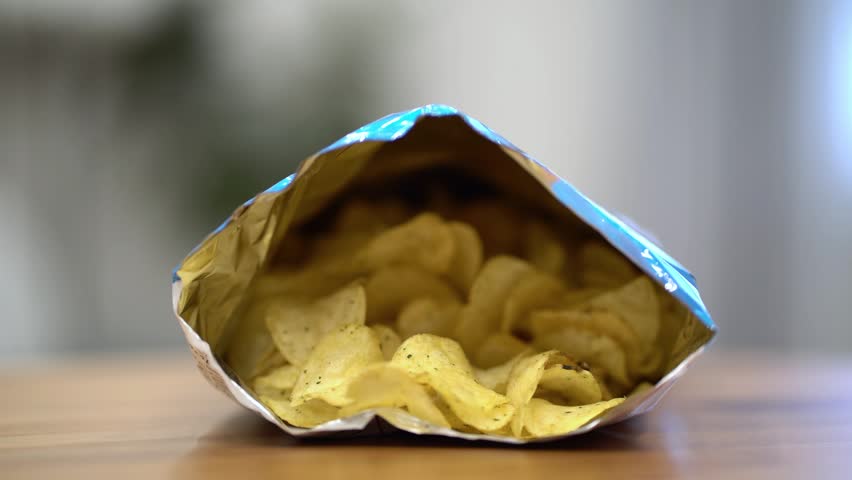 Close-up of female hands with red nails reaching into an open bag of golden nacho chips on a wooden table. Perfect stock footage for food, snacks, and lifestyle projects showing casual snacking and cravings.
