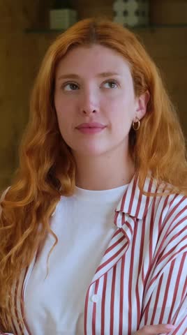 A beautiful young woman with striking red, wavy hair, dressed in a striped shirt, smiles and laughs, showcasing genuine joy and positive emotions. Her expressive face conveys happiness and a cheerful disposition.