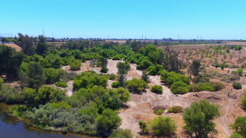 Reversing aerial drone footage pulling back over the San Joaquin River in Fresno, California, retreating along the waterway lined with lush riparian vegetation, trees, and bushes. Power lines and transmission towers are visible along the riverbank in the Central Valley landscape. This smooth, cinematic aerial pullback showcases the tranquil river, tree canopy, and scenic natural landscape from a serene birds eye perspective.