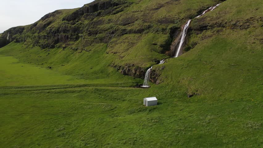 Aerial view of Gluggafoss waterfall cascading down green hills with a small shed in the foreground in Rangarthing eystra, Iceland.